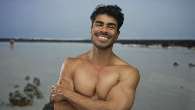 Man smiles as he crosses muscular arms over strong chest while standing on rocky beach shore under cloudy sky; confidence. - Powered by Adobe