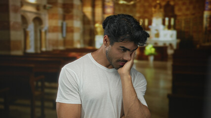 Man touching cheek and tilting head in church building with wooden pews and warm light; quiet reflection.