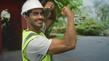 Man in white hardhat and neon vest smiles and flexes biceps in building under sunlight; confidence empowerment.