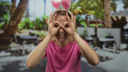 Woman wearing pink t-shirt and bunny ears headband making hand binoculars gesture on sunny...