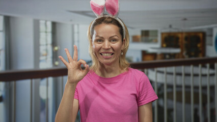 Woman making ok gesture with right hand in building lobby wearing bunny ears headband and bright...
