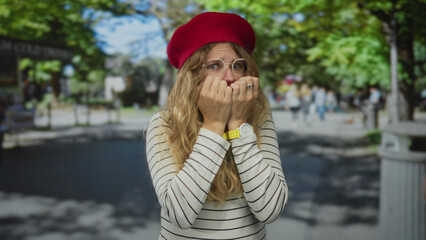 Woman brings hands to covering position in park over face looking anxious sporting blonde beret and...