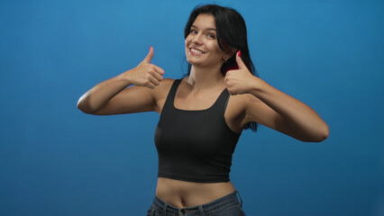 Woman wearing black crop top and denim jeans shows thumbs up gesture and smiles broadly in studio;...
