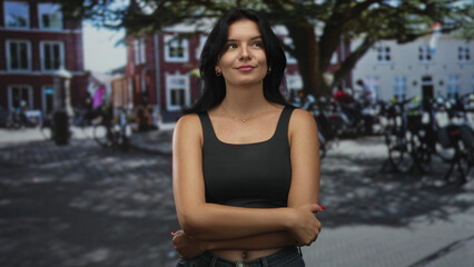 Young hispanic woman with arms crossed on sunny street wearing a black tank top and blue denim jeans; quiet confidence.