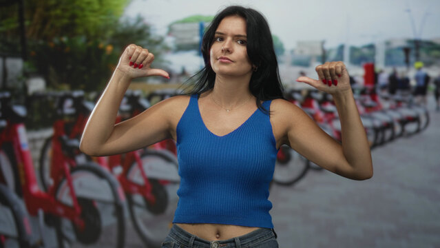 Fototapeta Hispanic young woman pointing both thumbs to herself beside a line of red rental bikes on a busy city street  pride.