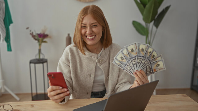 Woman smiling indoors holding phone and fan of us dollars next to laptop in cozy living room with plant in background.
