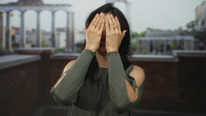 Woman smiling and covering face with hands in a green sweater at roman ruins building under soft...
