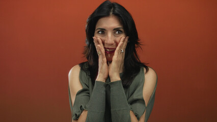 Hispanic middle aged woman with hands pressed against cheeks and wide open mouth in studio;...