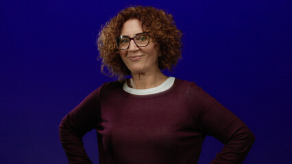 Woman with curly hair smiling confidently against an isolated blue background wearing glasses and...