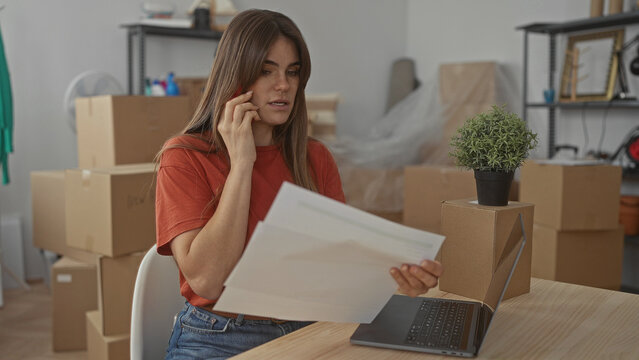 Woman talking on phone in living room surrounded by moving boxes reviewing documents with laptop on table in modern apartment setting. - Powered by Adobe