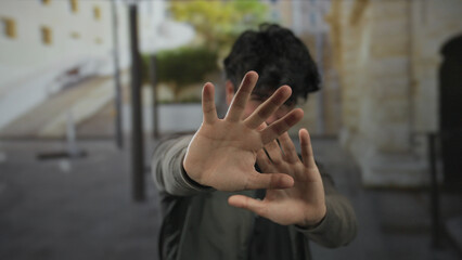 Young hispanic man with beard outdoors on sunny day makes stop gesture with hands in front of face showing rejection or refusal in urban setting with blurred background