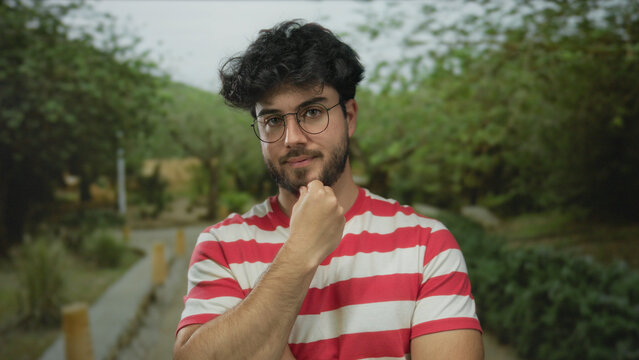 Young man with beard and glasses looking thoughtful in a park surrounded by lush greenery, wearing a striped shirt, embodying a pensive mood in an outdoor setting.