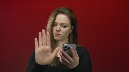 Woman raises palm and holds smartphone against red wall in studio with serious expression;...