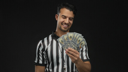 Young hispanic man referee holding us dollar bills with confidence against isolated black...