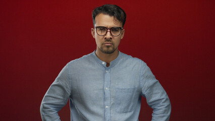 Fototapeta premium Young hispanic man in glasses and blue shirt pounds fist into palm against vivid red background, conveying anger and determination in isolated studio setting.