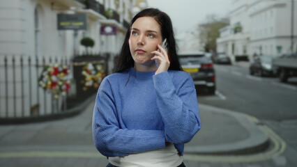 Fototapeta premium Woman on a phone call with serious expression stands on a city street wearing a blue sweater and dark pants, surrounded by urban scenery and parked cars on a cloudy day.