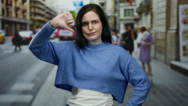 Woman expressing emotion with thumbs down gesture on busy outdoor urban street setting with blurred background of people and vehicles in motion.