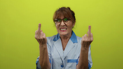 Elderly hispanic woman cleaner expressing defiance against a vibrant yellow backdrop while wearing...
