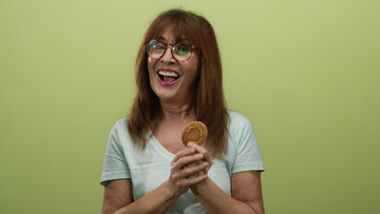 Senior hispanic woman with glasses holding a cookie against a yellow wall, smiling warmly while...