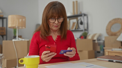 Hispanic woman using smartphone and credit card in new apartment living room surrounded by boxes suggests move or renovation, emanating focus and tech-savvy in cozy interior.