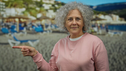 Woman with grey hair in pink sweatshirt points at beach background with blue sunbeds and umbrellas, smiling at seaside vacation scene showcasing relaxation and leisure vibes.