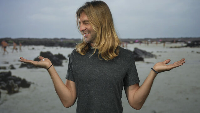 Man with long blond hair smiles softly and holds open palms on rocky coastline shore under cloudy sky; serenity.