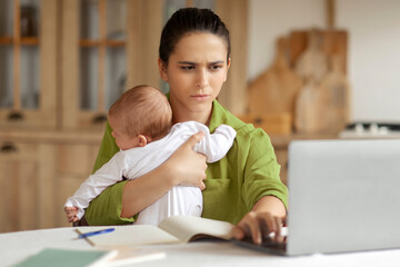 Tired mother tries to work with capricious infant child in her arms, typing on laptop computer...