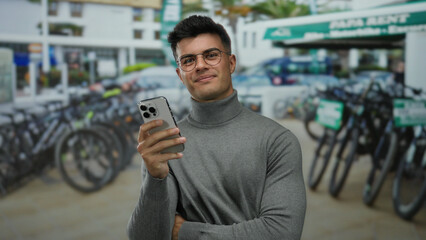 Young man in a turtleneck smiles while using his smartphone at a city bike rental station, framed...