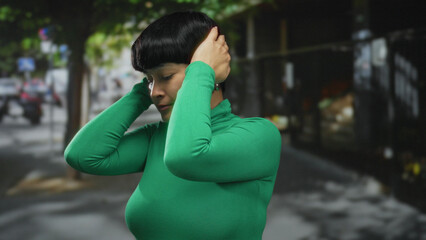 Woman in green sweater standing on city street covering her ears outdoors surrounded by urban scenery and green trees during daytime