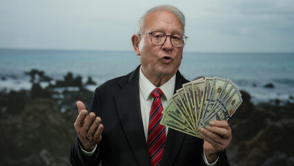 Senior man in business suit holding us dollars at seaside beach, conveying success and wealth with ocean backdrop.
