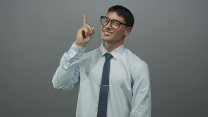 Man points finger upward in grey studio while wearing glasses and tie and smiling at ceiling;...
