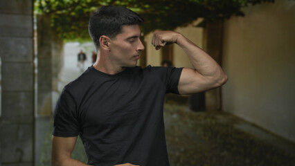 Young hispanic man flexes toned bicep and smiles on sunlit street under arched stone walkway;...