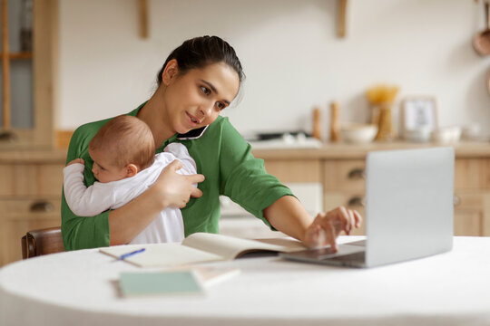 Busy mother caring baby having business phone call, sitting at table and using laptop. Remote job from home, babysitting and telecommuting