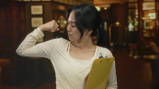 Young chinese woman in a hotel smiling confidently holding a yellow clipboard indoors, capturing a warm welcoming gesture in a professional setting. - Powered by Adobe