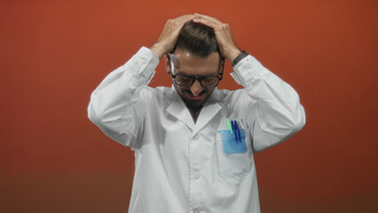Young hispanic scientist in lab coat grips head with both hands and clenches teeth in orange studio; stress.