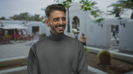 Fototapeta premium Hispanic man in gray turtleneck with beard smiling broadly under sunlight on busy street; optimism leisure.