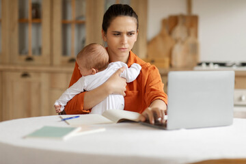 Tired mother tries to work with capricious infant child in her arms, typing on laptop computer...