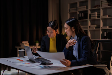 In a dimly lit office, business asian women collaborate late into the night. This image captures the intensity and focus of teamwork necessary for project success.
