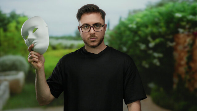 Young bearded man holding white mask in outdoor park setting with lush green trees visible in background creating an atmosphere of mystery and curiosity as he unveils his face to the camera