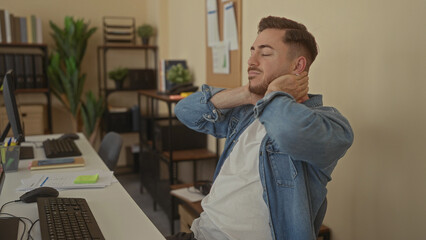 Fototapeta premium Young caucasian man with beard in office stretches neck wearing casual denim surrounded by plants and office equipment, creating a relaxed indoor work environment.