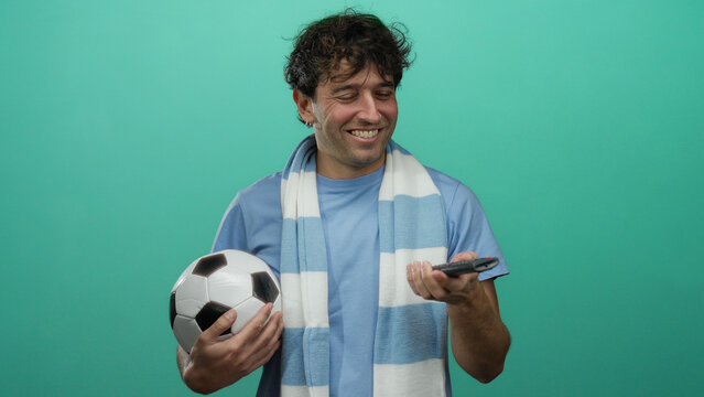 Hispanic man holding football and remote on green background, smiling enthusiastically, capturing the essence of sports fandom and leisure.