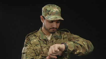 Young hispanic man in camouflage uniform points finger to watch with dogtags in studio; punctuality...