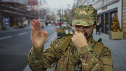 Man wearing camouflage uniform with backpack and dogtags pinches nose on busy city street; disgust.