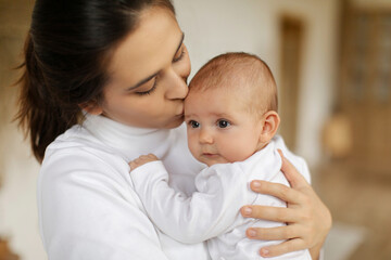 Caring mother holding and kissing her cute infant baby, expressing caress and tenderness, closeup shot. Happy motherhood and child care concept