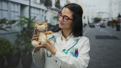 Young hispanic woman doctor with stethoscope holding stuffed toy lion in outdoor urban street setting conveying warmth and professionalism