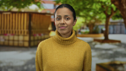 Confident young woman in a mustard sweater stands outdoors on a sunny street, surrounded by lush greenery and urban scenery, exuding a calm and approachable demeanor.