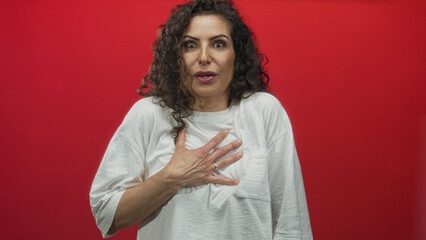 Young hispanic woman wearing white tshirt clutching chest with stunned expression in red studio;...