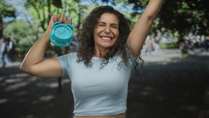 Woman raises arm and holds turquoise clock aloft in park while smiling; joy celebration awakening energy.