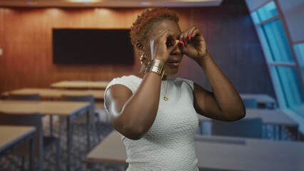 Young african american woman holds hands forming binoculars in classroom building; curiosity...