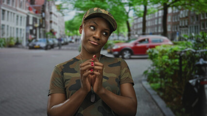 Woman in camouflage uniform clasps dogtags and folds hands on a street with parked cars and brick...
